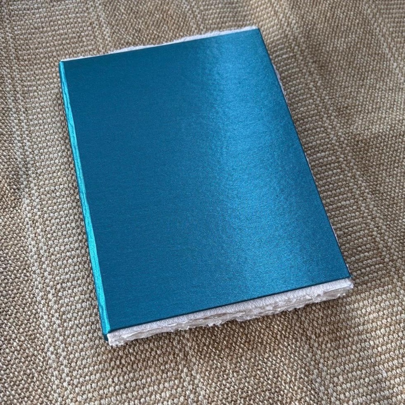 Op de afbeelding: Een turquoise notitieboek met een getextureerde kaft en witte, getextureerde randen. Het rechthoekige notitieboek ligt op een beige, geweven oppervlak. De kaft heeft een lichte glans.