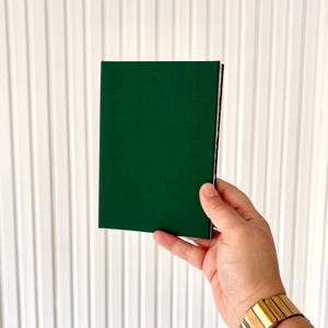 Puede incluir: Un peque&ntilde;o cuaderno verde oscuro con una cubierta verde lisa y un lomo negro. Se sostiene sobre un fondo blanco. La imagen est&aacute; bien iluminada, resaltando la textura de la cubierta.