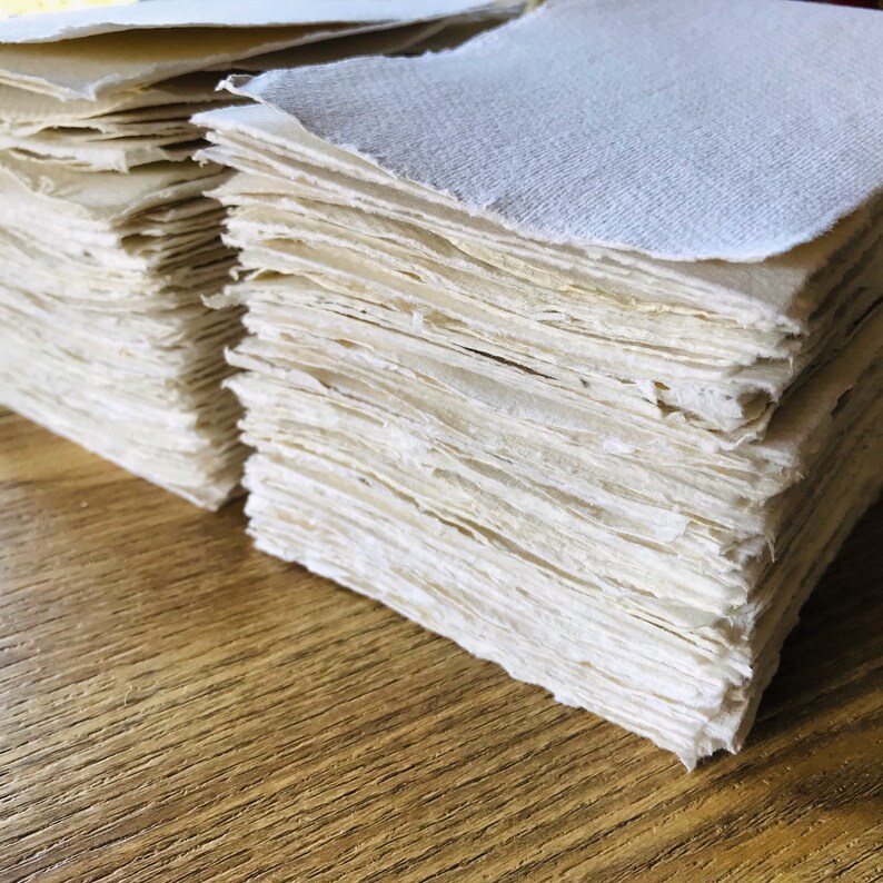 K&ouml;nnte beinhalten: Ein Stapel handgemachtes Papier, mit einer rauen, strukturierten Oberfl&auml;che in hellcreme. Das Papier liegt auf einer Holzoberfl&auml;che.