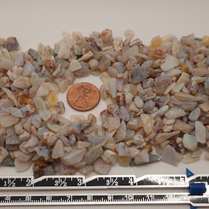 Puede incluir: Un montón de pequeños fragmentos de ópalo de forma irregular en tonos blancos, beiges y grises azulados. Una moneda de un centavo está en el centro para comparar el tamaño. Una regla muestra las medidas.