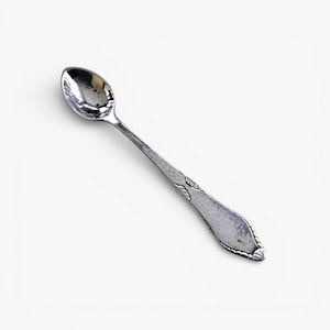 Può includere: Un cucchiaio d'argento con una piccola coppa ovale e un lungo manico. Il manico presenta un motivo decorativo a foglie vicino all'estremità. Il cucchiaio è posto su uno sfondo bianco.