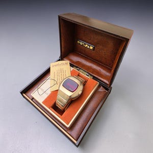 May include: A vintage gold-toned Pulsar digital watch with a red display, presented in its original brown box. The watch is displayed on a gold-colored base within the box, and a small card is visible. The box has the Pulsar logo.