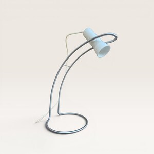 May include: A modern desk lamp with a white cylindrical shade and a curved, silver-colored metal frame. The base is a circular design, and the lamp has a white power cord. The lamp is designed in a minimalist style.