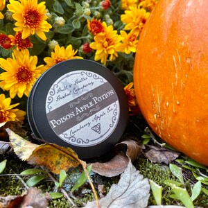May include: A black metal tin with a label that reads "Poison Apple Potion" with a cranberry and apple scent. The tin is surrounded by fall foliage and a pumpkin.