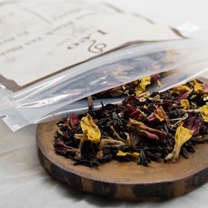 May include: A clear plastic bag with a label that reads "Loose Leaf Tea" is open and spilling out a blend of black tea, red and yellow flower petals onto a wooden surface.