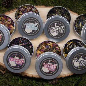 May include: Eight round metal tins with lids containing loose leaf tea blends. Each tin has a black and white label with a state outline and the name of the tea blend. The blends are Poppy, Hydrangea, Rose, Dahlia, Violet, Carnation, Sunflower.