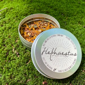 May include: A silver tin with a label that reads "Hephaestus god of fire and the forge" and "Spicy and Warming" with a hand-drawn illustration of a forge. The tin is filled with a blend of dried herbs and spices.