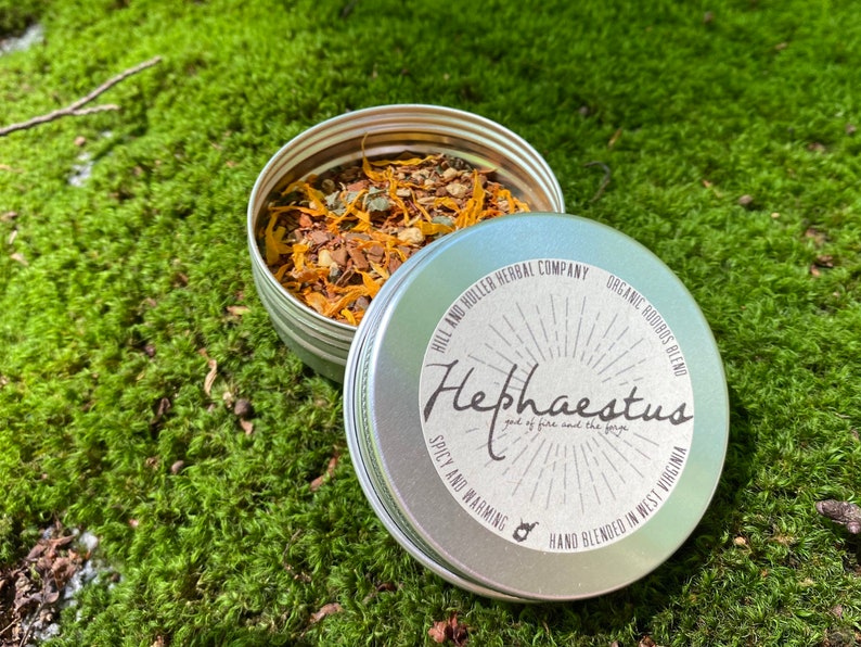 May include: A silver tin with a label that reads "Hephaestus god of fire and the forge" and "Spicy and Warming" with a hand-drawn illustration of a forge. The tin is filled with a blend of dried herbs and spices.