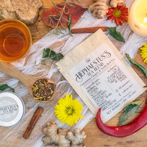 May include: A brown paper bag with a label that reads "Hephaestus's Tea Blend: Cinnamon, Ginger, & Nettle Red Tea". The bag contains a blend of loose leaf tea, including rooibos, nettle, cinnamon, ginger root, calendula, petals, black cohosh, rosemary, and dried cayenne. The bag is surrounded by various ingredients, including ginger, cinnamon sticks, red chilli peppers, and flowers.