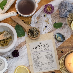 Puede incluir: Una bolsa de papel marrón con una etiqueta blanca que dice "HEKATE'S TEA BLEND Valerian & Elder Purple Tea" con una lista de ingredientes e instrucciones de preparación. La bolsa está rodeada de varias hierbas, flores y cristales.