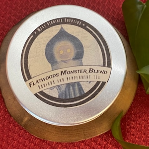 May include: A silver tin with a black and white label that reads "Flatwoods Monster Blend Rooibos and Peppermint Tea". The label features a cartoon illustration of a creature with glowing eyes.