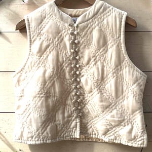 May include: Cream-coloured quilted vest with a diamond pattern and small button closures down the front. The vest is hanging on a wooden hanger against a light wood background, decorated with floral branches.
