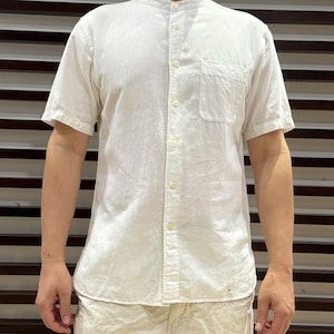 May include: A cream-coloured short-sleeved shirt with a mandarin collar and a single chest pocket. The shirt has a button-down front and is paired with matching cream-coloured trousers. The fabric appears lightweight, suitable for warmer weather.