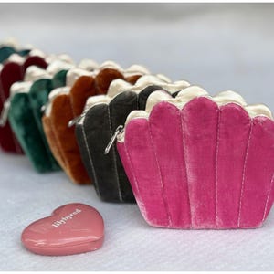May include: A row of velvet shell-shaped purses in shades like pink, green, and brown, each with a satin lining and a silver zip. A small, pink heart-shaped compact with the text "lilybyred" is in the foreground.