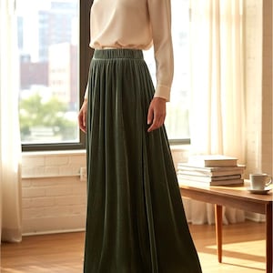 May include: A long, floor-length olive green velvet skirt with an elasticized waistband, paired with a long-sleeved, cream-colored silk blouse featuring a round neckline and buttoned cuffs, worn with matching olive green ankle boots.