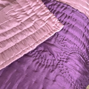 May include: A quilted textile featuring two distinct layers of fabric. The top layer is a soft light pink, while the bottom layer is a vibrant purple. Both sections are adorned with a consistent grid of small, visible running stitches.