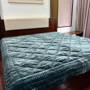 May include: A teal quilted bedspread with a diamond pattern on a bed. The bedspread is made of a soft, velvety material. The bed frame is dark wood, and a framed floral artwork hangs above the bed.
