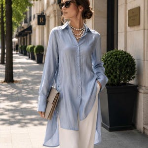 May include: A light blue button-down shirt with a high-low hemline, paired with white pants. The shirt has long sleeves and is worn with a pearl necklace and sunglasses. A beige clutch is held in the hand.
