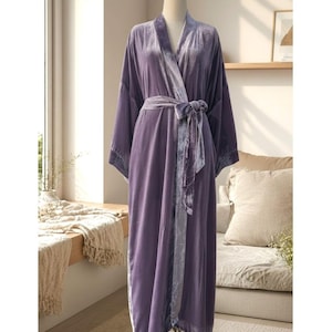 May include: A floor-length robe made of purple velvet fabric, featuring long sleeves, a shawl collar, and a matching tie belt at the waist. The garment has a smooth, reflective texture and is displayed on a mannequin to show its full length.