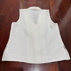 Puede incluir: Una blusa sin mangas de lino blanco roto con cuello y cuello en V. La blusa tiene aberturas laterales en el dobladillo y est&aacute; hecha de una tela ligera y texturizada. La prenda est&aacute; tendida sobre una superficie de madera oscura.