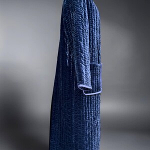 May include: A long, quilted robe in a deep navy blue color. The robe features a textured pattern, a collar, and a pocket. The edges are trimmed with a lighter blue. The robe is displayed on a mannequin.