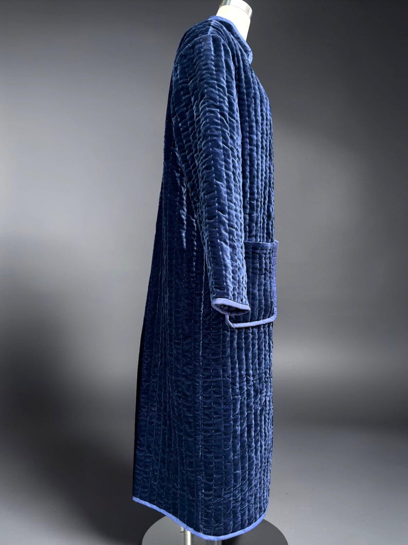 May include: A long, quilted robe in a deep navy blue color. The robe features a textured pattern, a collar, and a pocket. The edges are trimmed with a lighter blue. The robe is displayed on a mannequin.