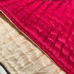 May include: A reversible quilted fabric piece featuring a vibrant magenta side with a grid-like stitched texture and a contrasting cream-colored side with a similar quilted pattern. The material displays a subtle sheen and visible horizontal stitching.