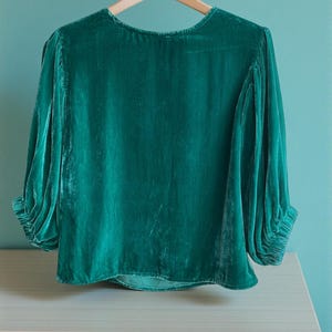 May include: A teal green velvet blouse with long sleeves and a round neckline. The sleeves are gathered at the cuffs. The blouse is hanging on a wooden hanger, resting on a light wood surface.