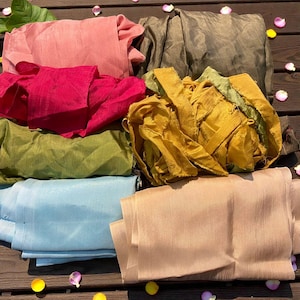 May include: Assortment of colorful silk fabric pieces in shades of pink, red, green, gold, blue, and tan. The fabrics are arranged on a wooden surface, showcasing their textures and colors. Ideal for sewing and crafting projects.