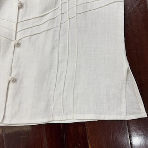 Puede incluir: Primer plano de una camisa de lino blanco con pliegues decorativos y cierres de botones. La camisa presenta una tela texturizada y un dise&ntilde;o a medida, lo que sugiere un enfoque en los detalles y la artesan&iacute;a. Los botones son de color blanco roto a juego.