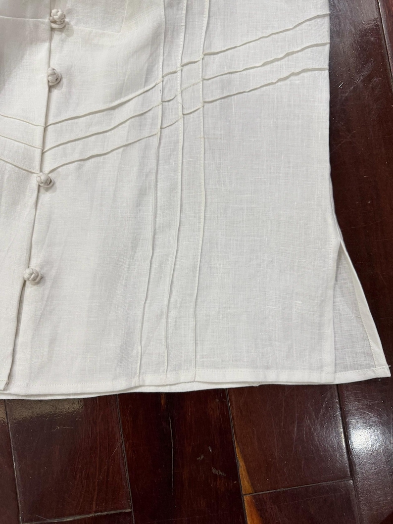 Puede incluir: Primer plano de una camisa de lino blanco con pliegues decorativos y cierres de botones. La camisa presenta una tela texturizada y un dise&ntilde;o a medida, lo que sugiere un enfoque en los detalles y la artesan&iacute;a. Los botones son de color blanco roto a juego.