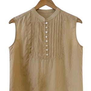 May include: A sleeveless, tan-colored blouse with a mandarin collar and a series of small, white buttons down the front. Vertical pleats adorn the center panel, adding texture. The blouse hangs on a wooden hanger against a white background.
