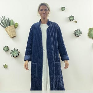 May include: A long, navy blue quilted robe with a mandarin collar and two front pockets. The robe is made of a soft, velvety fabric. The model is wearing a white dress underneath. The background features decorative succulents.
