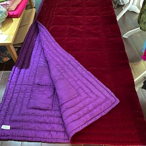 Könnte beinhalten: Eine wendbare, gesteppte Decke mit einer tiefroten Samtaußenseite und einer leuchtend violetten Innenseite. Die rechteckige Decke hat ein gestepptes Gittermuster, das ein luxuriöses und gemütliches Design zeigt. Sie liegt auf einem Holzboden.