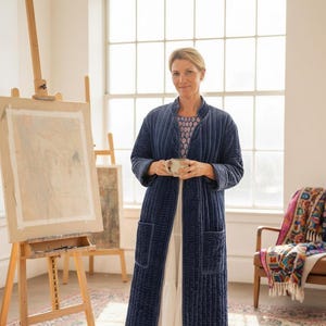 May include: A woman in a long, navy blue quilted robe stands in an artist's studio, holding a mug. The studio features easels with artwork, a patterned rug, and a chair with a colorful throw. The room is filled with natural light.