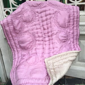 May include: A quilted blanket in a soft pink hue, featuring a repeating circular pattern. The blanket is made of a silky material and has a contrasting off-white underside. The stitching creates a textured surface.