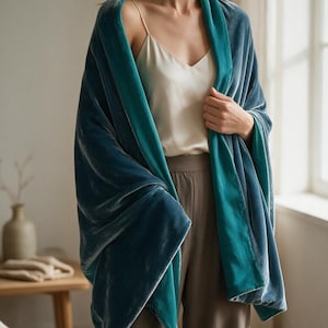 May include: A reversible velvet shawl featuring a deep slate blue exterior and a vibrant teal interior. The fabric has a soft, light-reflecting sheen, and the wrap is draped over the shoulders, showcasing the contrasting color layers of the material.
