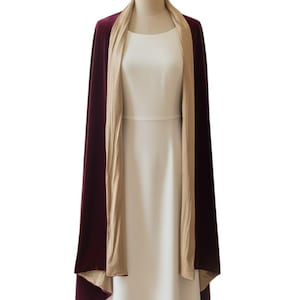 May include: A floor-length velvet wrap in a deep burgundy color featuring a contrasting beige lining. The wrap is draped over a plain white dress on a mannequin, showcasing a smooth texture and a long, open-front design that hangs straight down.