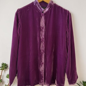 Peut inclure: Une chemise à manches longues en velours violet profond dotée d'un col officier et d'une fermeture boutonnée sur le devant avec des boutons recouverts de tissu assorti. Le vêtement présente une texture douce et réfléchissante sur un cintre.
