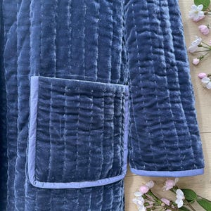May include: A quilted, navy blue velvet robe with a light blue trim around the pocket and cuffs. The robe has vertical stitching and a soft, luxurious appearance. The image shows a close-up of the robe's texture and design.