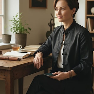May include: A person seated at a wooden desk, wearing a dark grey jacket with black buttons over a white top, and black trousers. They are holding a smartphone. A cup of tea, a notebook, and a pen are on the desk.