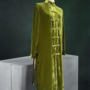 Può includere: Lungo cappotto in velluto verde oliva con collo alla coreana e chiusure a ranella. Il cappotto ha maniche lunghe e uno spacco laterale alto. L'indumento è esposto su un manichino.