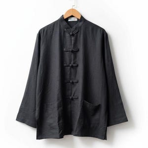 Könnte beinhalten: Ein dunkelgraues Langarmhemd mit Stehkragen und Froschverschlüssen. Das Hemd hat zwei Vordertaschen und hängt an einem Holzbügel. Der Stoff scheint Leinen oder ein ähnliches leichtes Material zu sein.