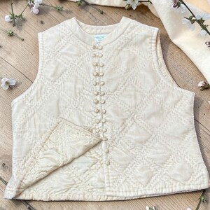 May include: Cream quilted vest with a diamond pattern and a row of small buttons. The vest has a rounded neckline and a small pocket. The background includes flowers and a cream-coloured cloth.
