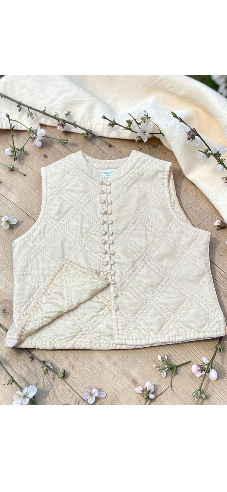 May include: Cream quilted vest with a diamond pattern and a row of small buttons. The vest has a rounded neckline and a small pocket. The background includes flowers and a cream-coloured cloth.