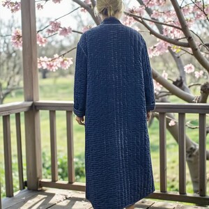 May include: A long, quilted navy blue robe with a vertical stitched pattern. The robe has long sleeves and a collar. The person is wearing the robe on a wooden porch with a view of a flowering tree.