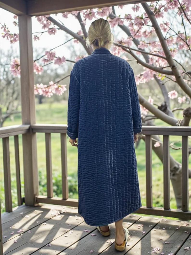 May include: A long, quilted navy blue robe with a vertical stitched pattern. The robe has long sleeves and a collar. The person is wearing the robe on a wooden porch with a view of a flowering tree.