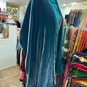 May include: A teal velvet wrap or shawl featuring a smooth, reflective texture and a wide, structured collar in a slightly darker shade of teal. The garment drapes loosely, showcasing a soft sheen across the fabric surface and clean edges.