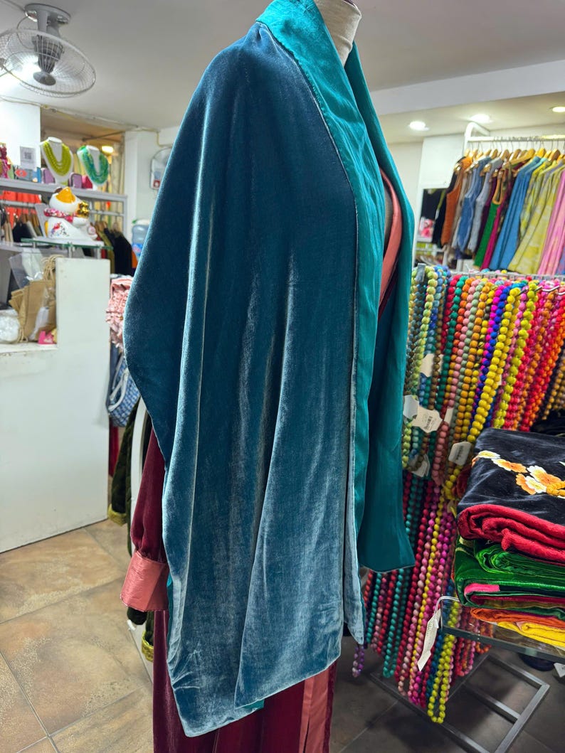 May include: A teal velvet wrap or shawl featuring a smooth, reflective texture and a wide, structured collar in a slightly darker shade of teal. The garment drapes loosely, showcasing a soft sheen across the fabric surface and clean edges.