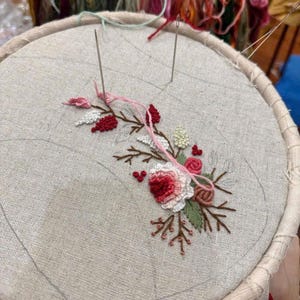 Puede incluir: Primer plano de un dise&ntilde;o de bordado floral en un trozo de tela blanca estirada en un aro de bordado de madera. El dise&ntilde;o presenta flores rosas y rojas con hojas verdes.